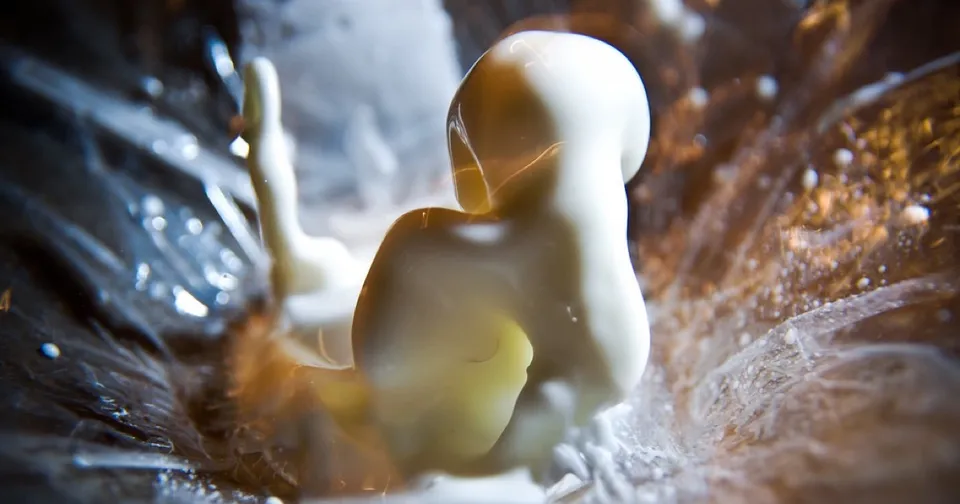 Macro photograph of a white non-Newtonian fluid folding and twisting against a dark reflective surface, evoking the fluid instabilities at the heart of the Navier-Stokes blowup problem.