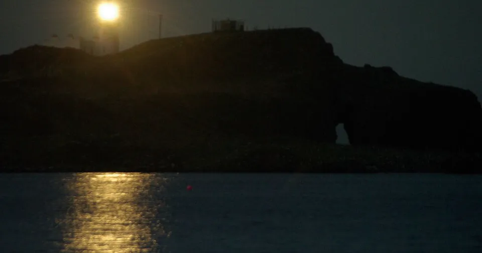 The lighthouse on Fidra lit against a dark night sky, its lamp reflected in the sea below, standing in for an agent that must hold its course across weeks of changing conditions.
